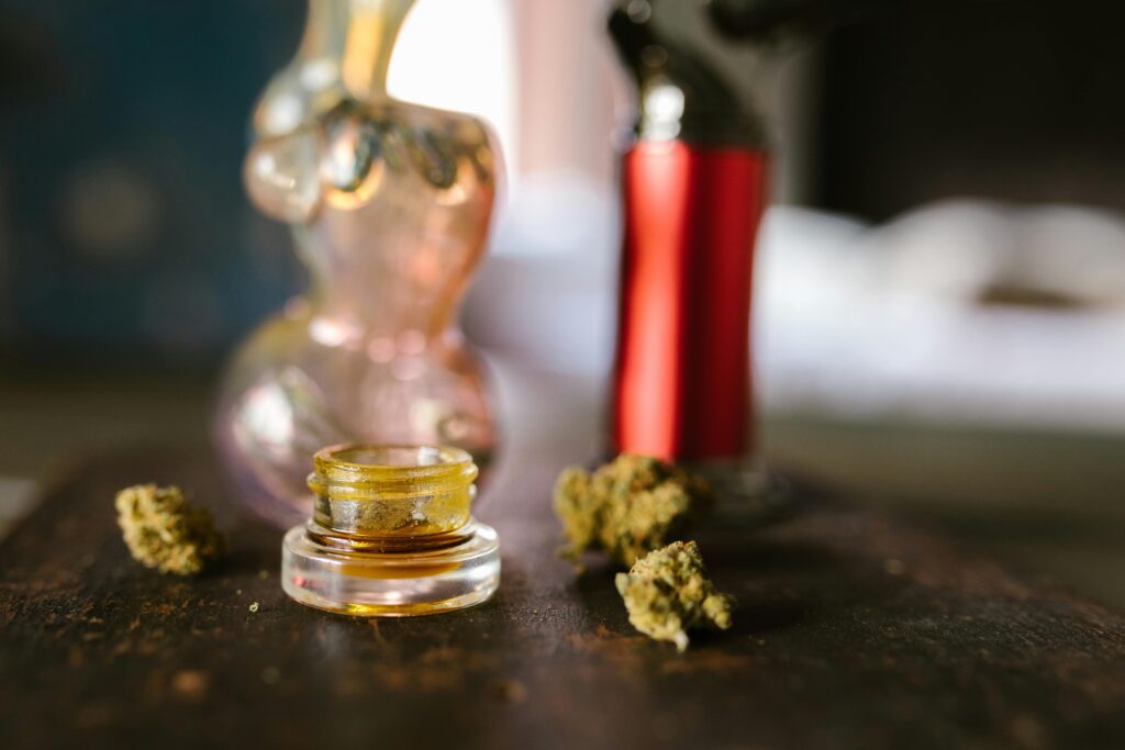 Cannabis concentrate in a jar and loose cannabis flower sit on a table with a bubbler and mini torch behind them.